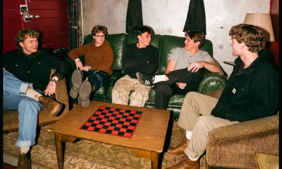 Bandmates from The Band Solstice sit on couches and pose in front of a brown table with a checkerboard. 