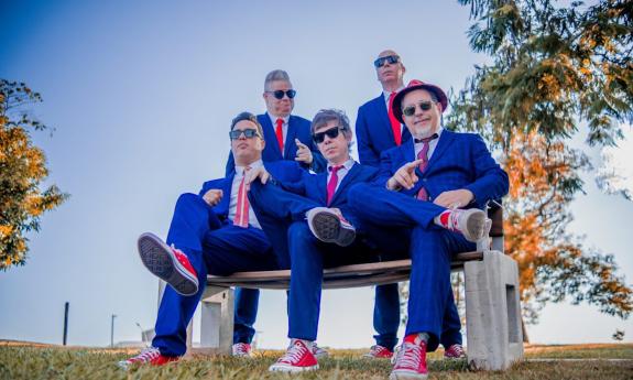 The Blues Beatles, dressed in blue suits and matching ties, sit together on a small bench for a posed shot.