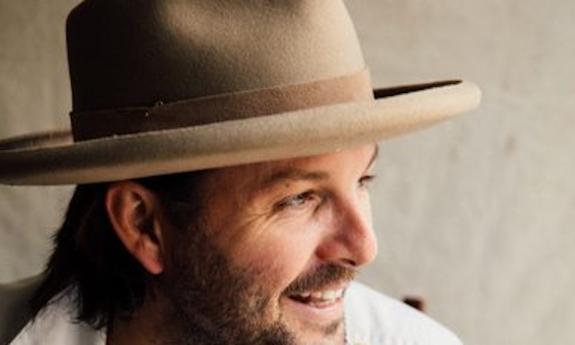 Keith Harkin wears a brown wide-brimmed hat and smiles at an object in the distance.