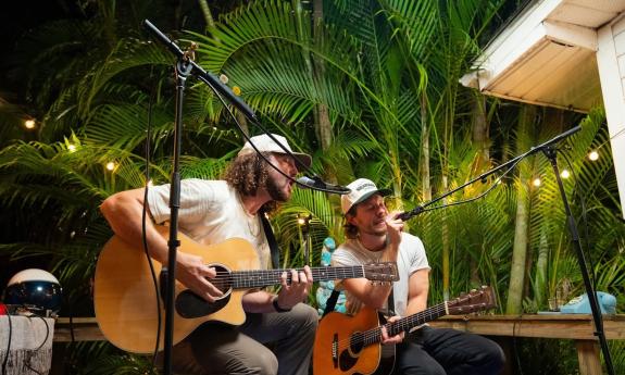 Matt and Zack strum their guitars on stage against a backdrop of lush leaves and bushes.
