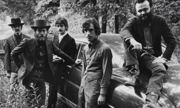 The Band’s members, dressed in dark attire, cluster together beside a vintage car.