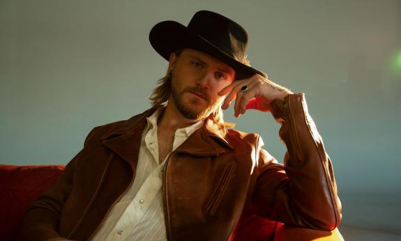 Musican Jackson Dean, leaning on his arm, outside, wearing a cowboy hat and leather jacket