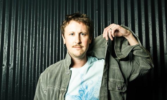 Johnny Pemberton grabs his green shirt and poses in front of a dark green backdrop.