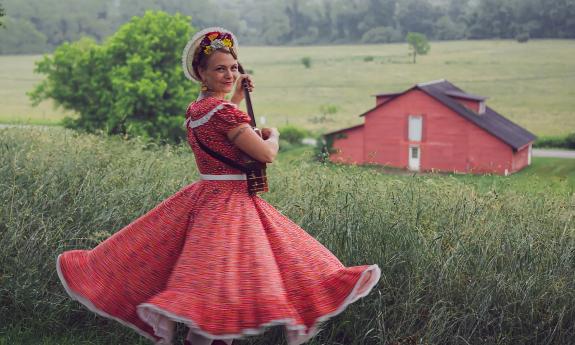 Sparrow Smith in a red country dress with petticats, in a green field with a red barn in the distance