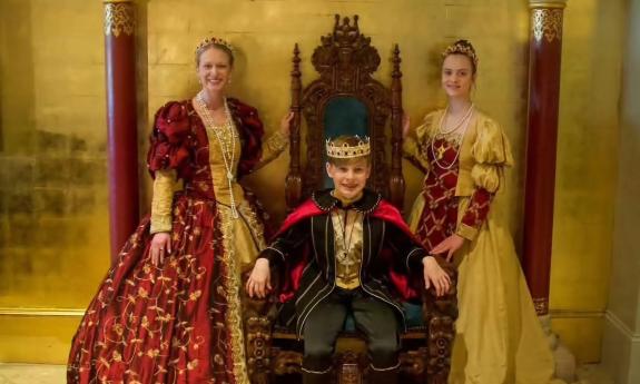 Two women in Spanish colonial garb, standing on either side of a young man dressed as the Spanish King