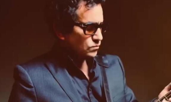A.J. Croce wears dark sunglasses and poses in front of a dark brown backdrop.