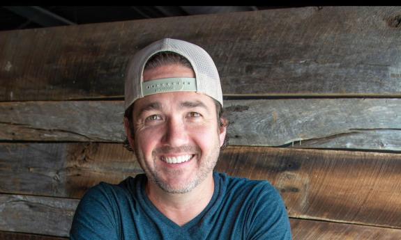 Songwriter Chris Blair in backwards ball cap, in front of a wooden wall