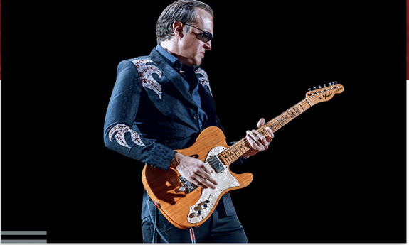 Joe Bonamassa strums his guitar in front of a black backdrop.