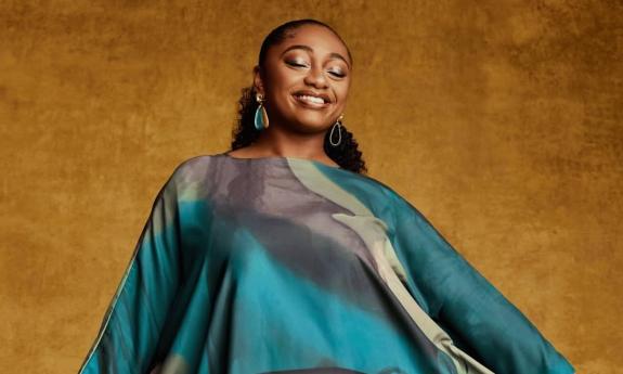 Samara Joy wears a flowing dress and smiles proudly in front of a gold backdrop.