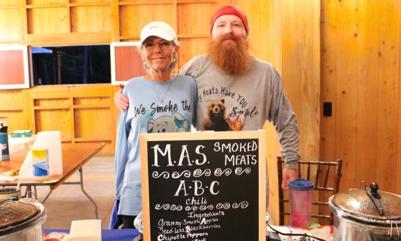 A man and a woman standing behind their chili cook-off table in a barn