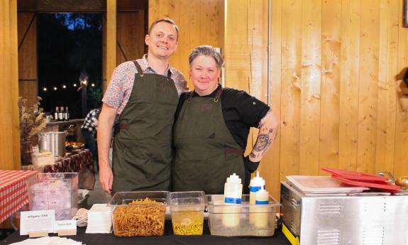 Two chefs in matching aprons standing at their table offering alligator chili