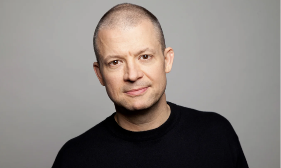 Jim Norton smiles and poses in front of a grey backdrop.