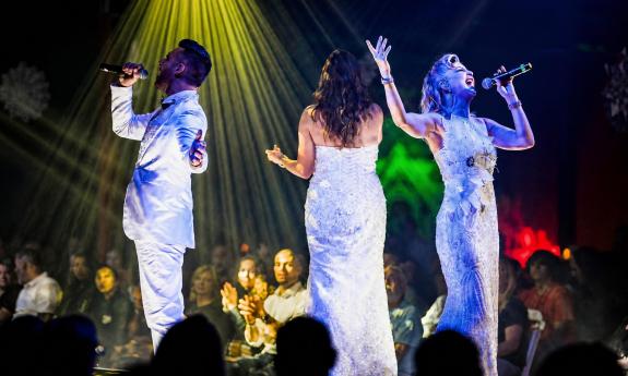 Three singers, all in white on stage with vibrant lighting