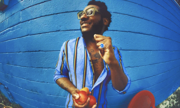 Leon Bridges wears a blue top and smiles while standing in front of a blue backdrop.