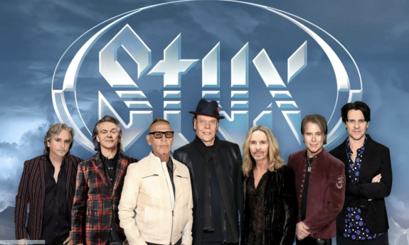 Bandmates from Styx huddle together and pose in front of a backdrop with their logo.