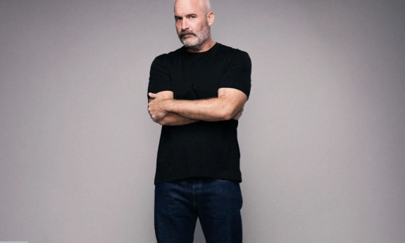 Tom Segura dons black clothing and poses in front of a grey backdrop.