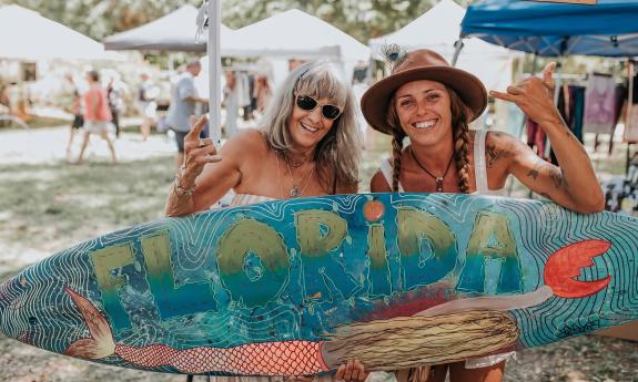 Two women holding a surfboard painted with a Florida motif