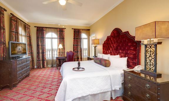 An opulent hotel room at the Casa Monica Hotel. A large bed with a red velvet headboard is in the center of the room, next to several lamps, a bedside table, and a television.