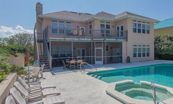 A vacation rental property in St. Augustine on a summer day. A pool and hot tub are in the foreground and the pink two story house is in the background. 