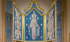 The triptych by Hugo Ohlms which honors and celebrates the Virgin Mary in the Cathedral Basilica of Saint Augustine in St. Augustine, Florida. Painted with royal blue and shining gold leafing, Mary occupies the center whilst angels flank her on either side. Saint Augustine, Florida is also the location of the first Marian shrine in the United States.