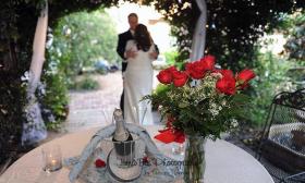 A bride and groom embrace under a trellis of jasmine at the Casa de Solana. Red roses and a bottle of champagne wait for them on table with a white tablecloth