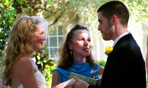 A bride and groom, sharing their vows under a tree during a sunny day, with an officient wearing blue assisting them