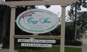 An ovular sign reading Cozy Inn: Daily & Extended Stay. An illustration of flowers and vines surrounds the cursive logo. Rectangular signs hang below
