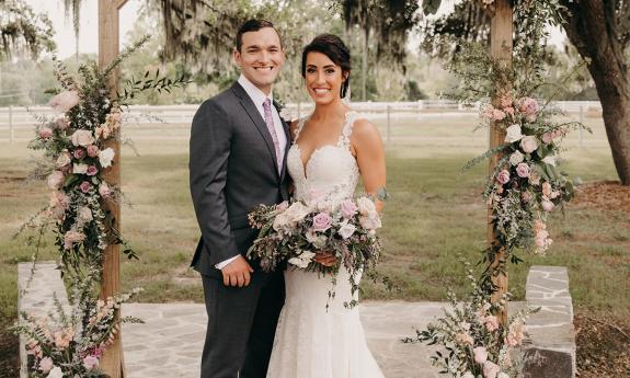 A couple stands on their wedding day with floral arrangements and bouquet by Feather + Bloom.