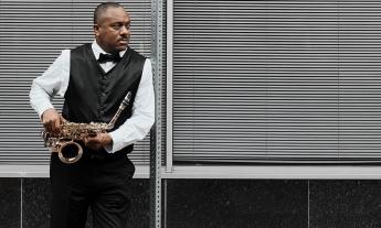 Marcus Click wears a black and white suit and holds onto his saxophone while posing for the camera