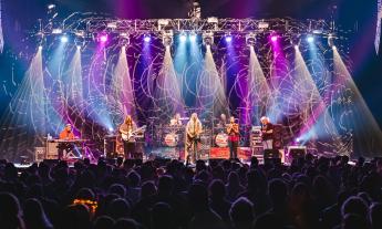 Band members of the Dark Orchestra performs live on a stage with colorful lights. 