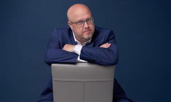 Comedian Danny Johnson poses in a blue blazer and glasses while sitting in a grey chair. 