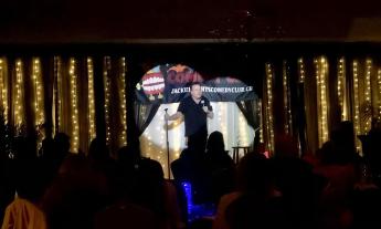 A male comedian talks to the audience, while standing on a spotlit a stage in front of a gold curtain