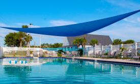 This large hotel pool has a bright blue triangular sun cover, deck chairs, and a white privacy fence.