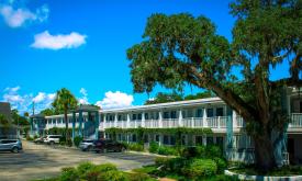 A two-story motel in the day, with bright blue sky, fluffy clouds, and a live oak tree.