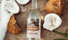 A bottle of Coconut Rum by City Gate Sprits, lying on a beach with open coconuts and froth from a wave