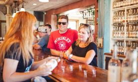 A woman Tasting Guide with long blond hair pouring sample drinks for a tourist couple in t-shirts and sunglasses