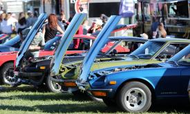 A line of four classic sport cars, red, black, green, and blue, with hoods open at an auto show