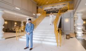 Groom in blue suit, standing at the foot of a staircase, back to the bride who is coming down the stairs