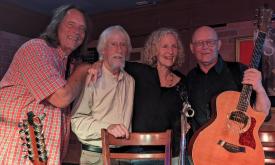 Four musicians, David Watt Beasley, Bob Patterson, Katherine Archer, and John Dickey stand together on stage with John holding a guitar