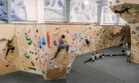 A wide photo of an indoor climbing facility with a number of people on the walls and others watching