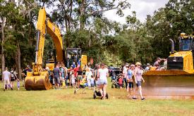 A lawn, with trees in the background, and a backhoe and bulldozer, with families viewing and touching the trucks