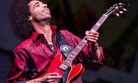 Selwyn Birchwood holds his guitar and smiles in front of a black backdrop. 