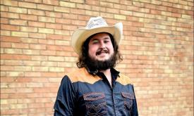 Lane Whalin, in western shirt and cowboy hat, against a yellow brick wall