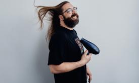 Gabriel Rutledge smiles while using a hair dryer in front of a gray backdrop. 