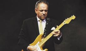 Jimmie Vaughan closes his eyes and strums his guitar in front of a black backdrop. 