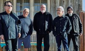 The members of Los Lobos pose in black attire against the rustic backdrop of an old house.