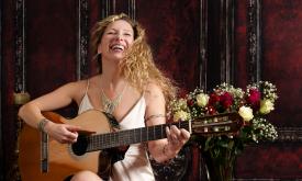 Zaza in a white dress, holding her guitar and singing.