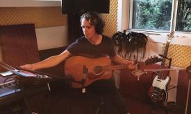 A musician seated in th ecorner of a living room, with musical equipment in an arc around him