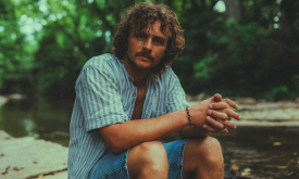 Evan Honer poses in front of a rocky lakeside wearing an open blue shirt with stripes. 