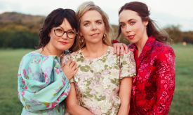 The members of I'm With Her stand together in floral tops, framed by a softly blurred green grove in the background.
